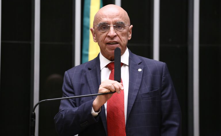 Deputado Mário Heringer fala ao microfone. Ele é um homem claro, calvo, usa um terno azul-marinho, camisa branca e gravata vermelha. No fundo, à direita, há uma bandeira do Brasil